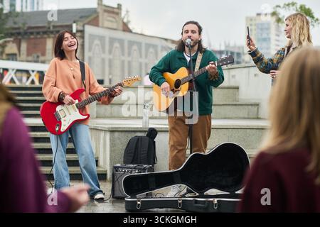 Young Caucasian woman playing electric guitar and singing with man playing acoustic guitar and singing outdoors while another woman recording performance on smartphone Stock Photo