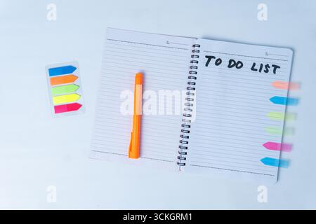 Flat lay of an open spiral notebook with handwritten to do list, orange pen, and colorful sticky notes on white background. Stock Photo
