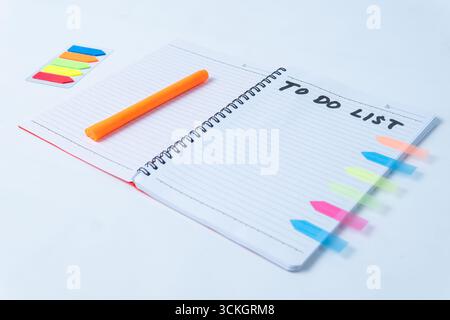 Flat lay of an open spiral notebook with handwritten to do list, orange pen, and colorful sticky notes on white background. Stock Photo