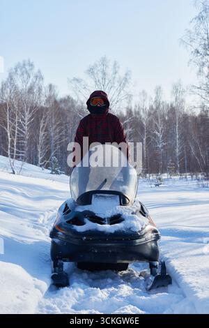 Young adult man riding snowmobile through snowy forest landscape, wearing winter gear and protective goggles, standing upright behind vehicle in outdoor winter setting Stock Photo