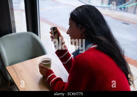 Smiling woman with long dark hair holding smartphone during video call in cozy cafe, with coffee in hand. Digital connection, relaxed mood and modern Stock Photo