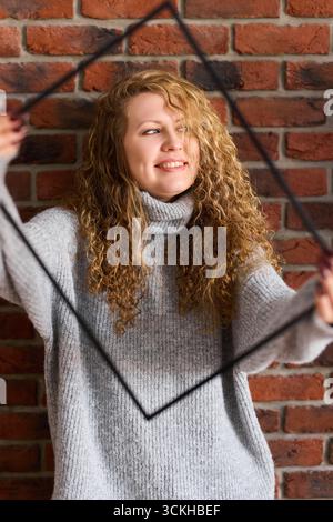 Young woman smiles while holding a frame in front of a brick wall Stock Photo