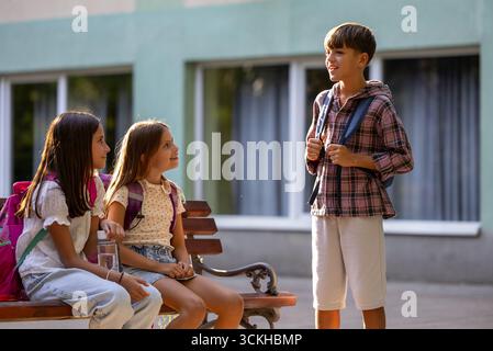 Three children relax on bench after school, engaging in lively conversation while enjoying the warmth of afternoon sun Stock Photo