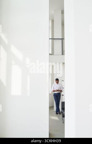 Man standing in modern bright office using smartphone, leaning against wall, focused on screen, natural light streaming through large windows Stock Photo