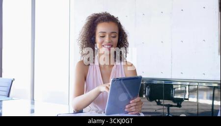 Smiling worker wearing blouse tapping tablet at glass desk in modern office, with wireless headset Stock Photo