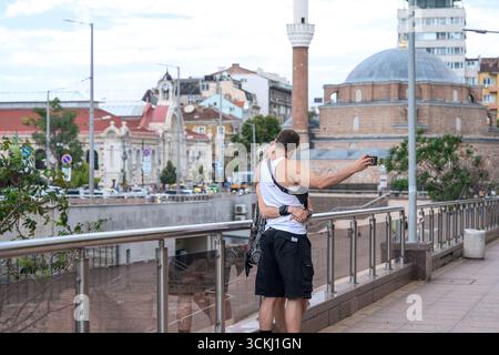 A couple kissing while taking a selfie with the Banya Bashi Mosque and Central Sofia Market Hall in the background, Sofia, Bulgaria. Stock Photo