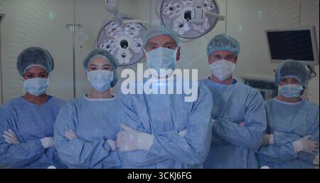 Five staff wearing gowns and masks standing in operating room, with overhead lights and monitor Stock Photo