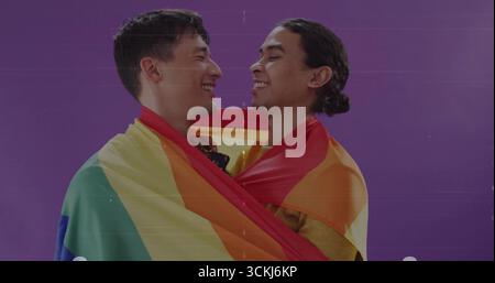 Embracing couple sharing smiles in studio against purple backdrop, wrapped in rainbow pride flag Stock Photo