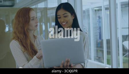 Reviewing laptop, colleagues wearing office attire working in corridor, with glass walls and lights Stock Photo