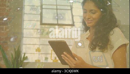 Smiling Indian student using tablet in office with potted plant, lamp, floating icons, copy space Stock Photo
