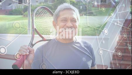 Smiling senior Hispanic man holding tennis racket over shoulder on backyard patio, network overlay Stock Photo