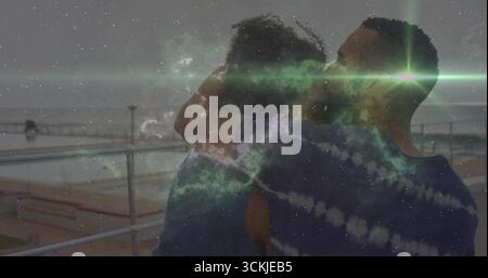Embracing woman and man in blue tops on boardwalk overlooking ocean with cosmic nebula overlay Stock Photo