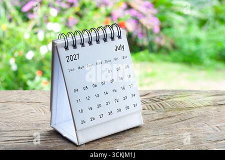 July 2027 spiral desk calendar on rustic wooden table with blurred green garden background. Stock Photo