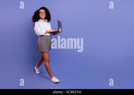 Young professional woman using a laptop while walking against a solid purple background Stock Photo
