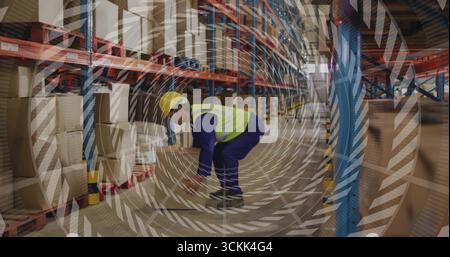 Kneeling worker wearing hard hat, safety vest lifting cardboard box on pallets in warehouse aisle Stock Photo