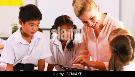 Kneeling teacher pointing tablet screen at students in uniforms in classroom with yellow poster Stock Photo