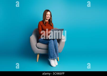 Smiling woman on chair using laptop in front of blue background, showcasing casual style and technology-focused lifestyle Stock Photo