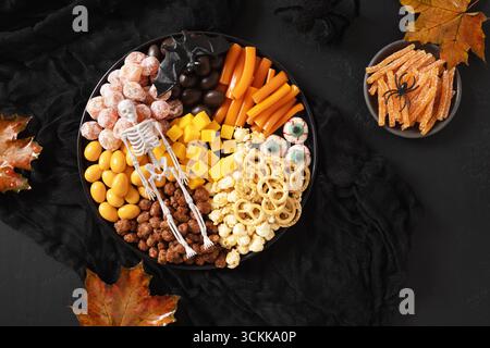 Halloween funny charcuterie board with sliced pumpkins, chocolate candy, spider, crackers on black background. View from above. Stock Photo