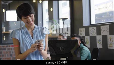 Chinese office worker in blue blouse tapping smartphone screen in office, with desktop monitors Stock Photo