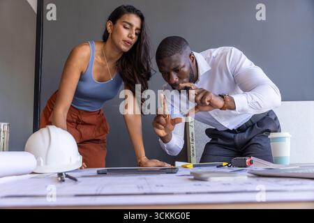 Diverse coworkers collaborating over architectural plans at office desk with blueprints, helmet Stock Photo