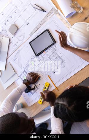 Diverse coworkers examining blueprints at drafting table with digital tablet, spirit level Stock Photo