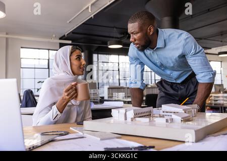Diverse coworkers discussing architectural model beside laptop in open-plan office with coffee Stock Photo