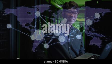 Standing IT professional in dark shirt holding laptop in green server room, with world map overlay Stock Photo