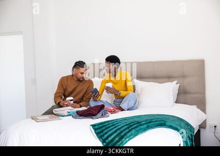 Diverse couple sitting on bed holding coffee cups while showing smartphone beside teal blanket Stock Photo