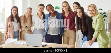 Smiling diverse women standing by desk in office hugging and laughing, showing unity and teamwork. Stock Photo