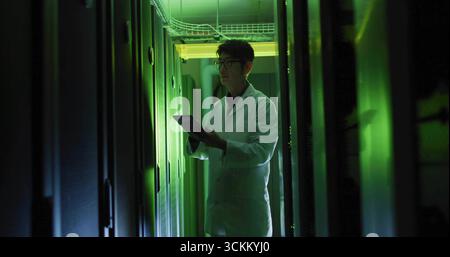 Reviewing tablet, Korean technician in lab coat inspecting racks with green lights in server room Stock Photo