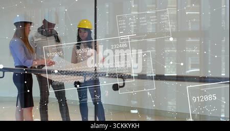 Reviewing architects wearing safety helmets examining blueprints at glass railing, with data panels Stock Photo