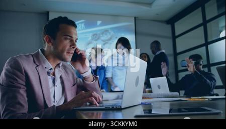 Man in blazer talking on smartphone while typing on laptop in meeting room, with projection screen Stock Photo