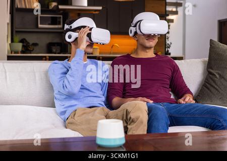 Male friends sitting on light sofa in living room using vr headsets with smart speaker Stock Photo