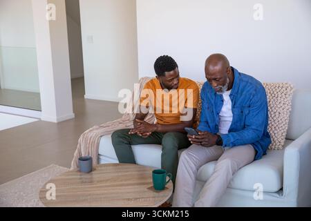 African american father and son on sofa in living room viewing smartphone near coffee mugs Stock Photo