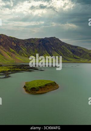 Serene aerial view of a grassy island in turquoise waters, surrounded by rugged volcanic mountains with sun rays breaking through the clouds. Stock Photo