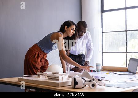 Diverse coworkers collaborating on architectural model at office desk using laptop, blueprints Stock Photo