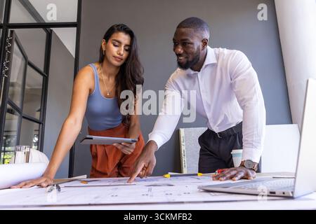 Diverse coworkers in business attire examining blueprints on table in office with tablet Stock Photo