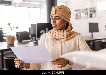Indian woman in tan turban-style headscarf reading white paper at open space with monitors Stock Photo