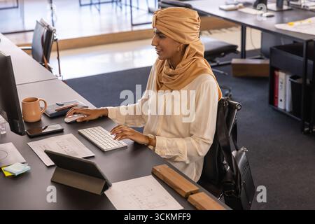 Asian indian woman sitting at open-plan office desk reviewing floor plans on tablet, keyboard Stock Photo