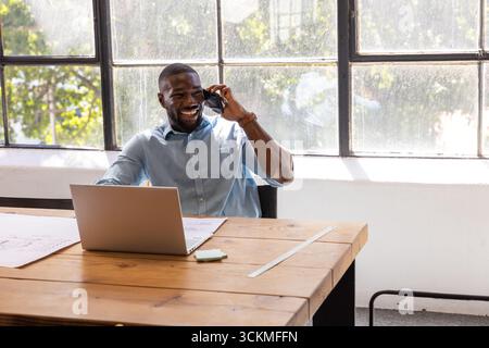 African American man smiling while talking on smartphone, reviewing laptop and plans at office desk Stock Photo