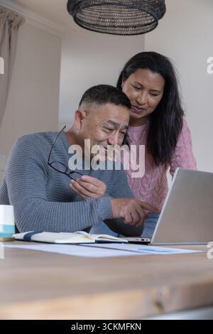 Diverse senior couple smiling, pointing at laptop at home on dining table with eyeglasses Stock Photo