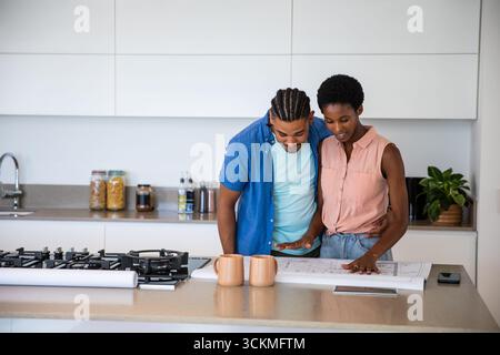 African American couple leaning over rolled-out blueprint on kitchen island, consulting smartphone Stock Photo