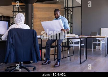 Diverse coworkers collaborating on blueprint sheet while examining computer in office Stock Photo