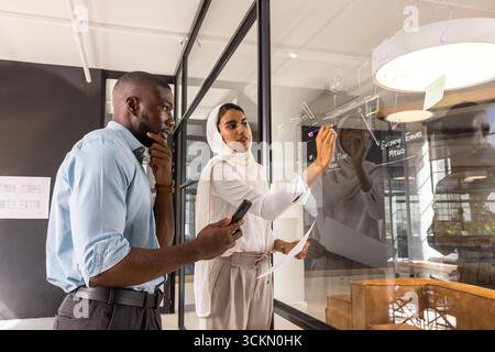 Diverse coworkers sketching designs on glass whiteboard at office while holding smartphone Stock Photo