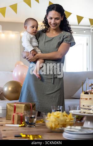 Diverse mother holding infant son in living room with cake on wooden table, balloons, gifts Stock Photo