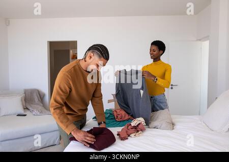 Diverse couple folding sweaters and tops on bed in bedroom with sofa, smartphone and cardboard box Stock Photo