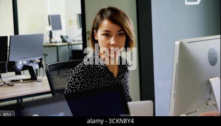 Typing female office worker focusing on screen at office desk, with laptop, glass partition Stock Photo