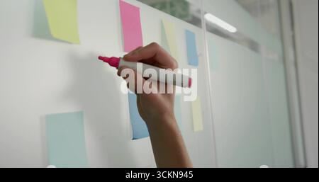 Holding hand gripping pink marker poised on glass board in office with pastel notes, copy space Stock Photo
