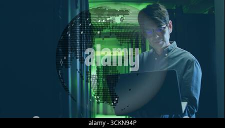 IT technician examining laptop screen in server rack corridor, with floating digital globe Stock Photo