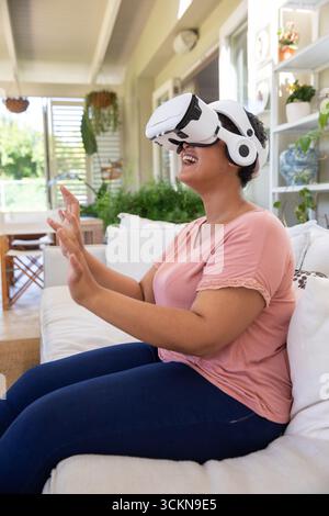 Woman wearing vr headset, reaching forward on sofa in sunroom with hanging planters, copy space Stock Photo